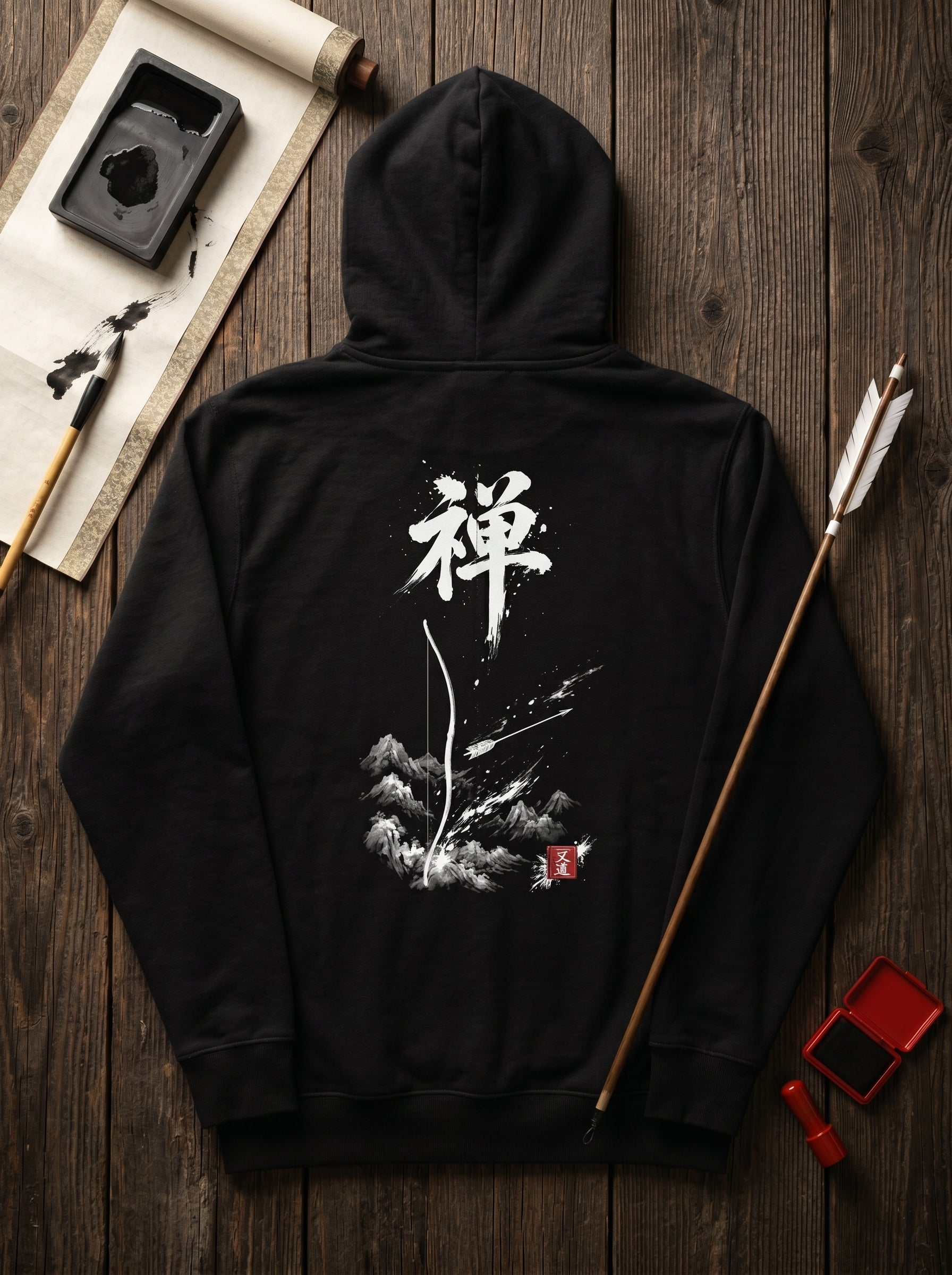 Flat lay of black hoodie back with Zen 禅 Sumi-e calligraphy and bow-and-arrow scene, surrounded by Japanese ink stone, calligraphy brush, Kyūdō arrows and red ink seal on wood surface, MINDŌ Studio
